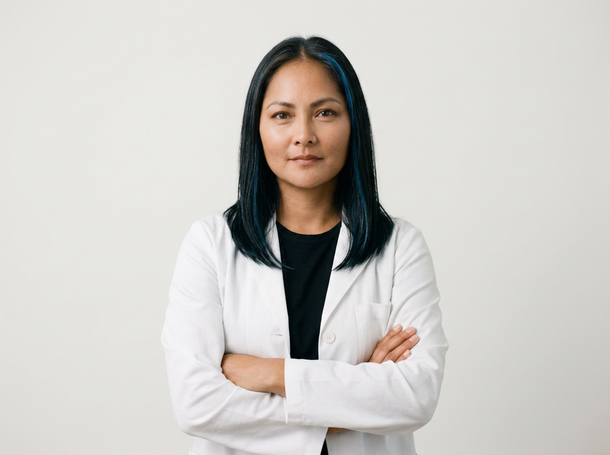 A woman with straight black hair and blue highlights, wearing a white lab coat over a black shirt, standing with arms crossed against a plain white background.