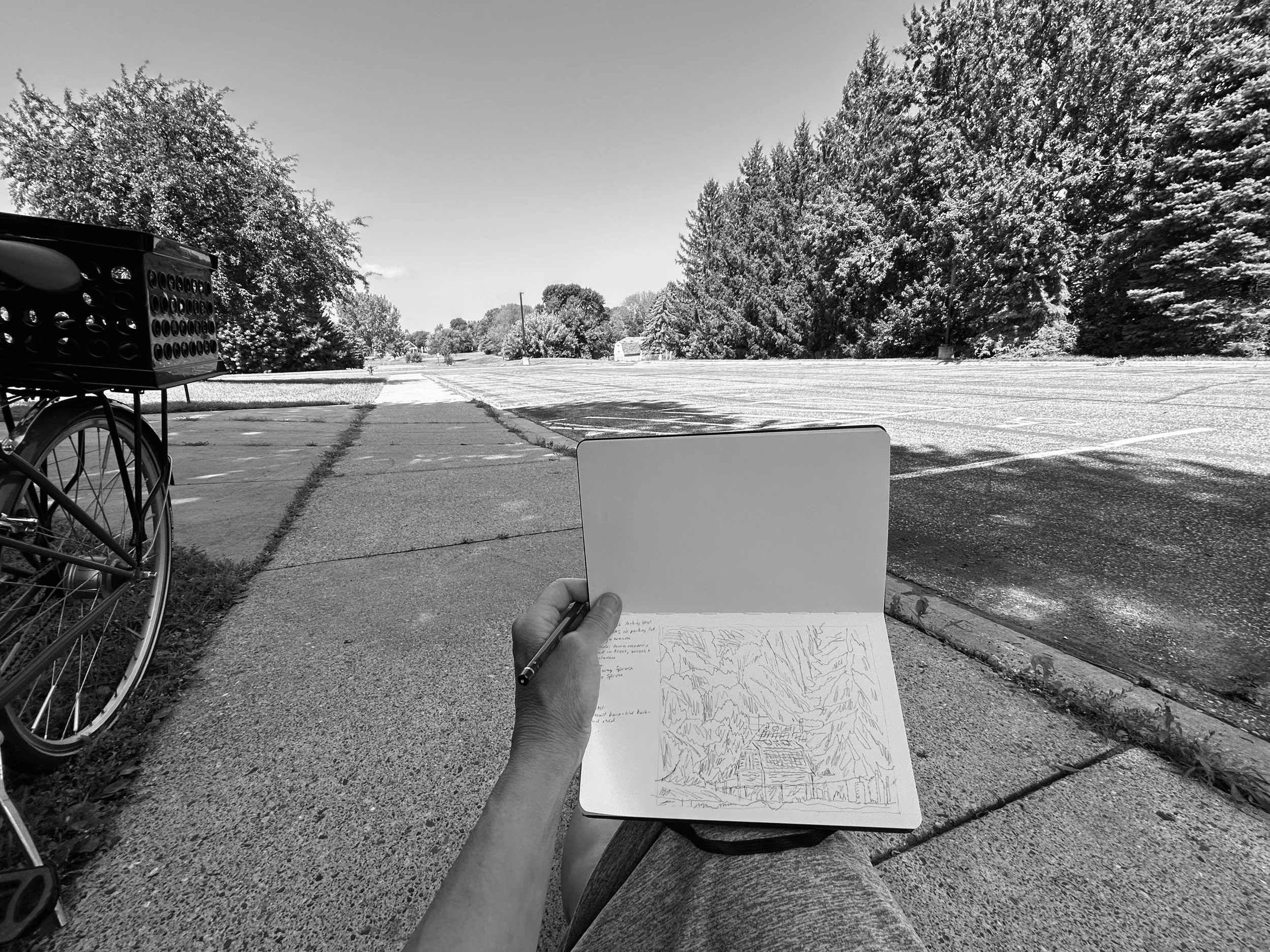 A photo of Michon Weeks holding a sketchbook with drawings of trees and landscapes, sitting outdoors near a sidewalk, with a parking lot, bicycle, and large trees in the background, all in black and white.