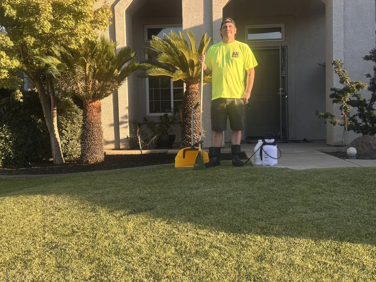 Man standing on front lawn with poop scooping tools and kennel grade disinfectant sprayer, wearing bright yellow T-shirt and shorts, next to a house with clean lawn and trees.