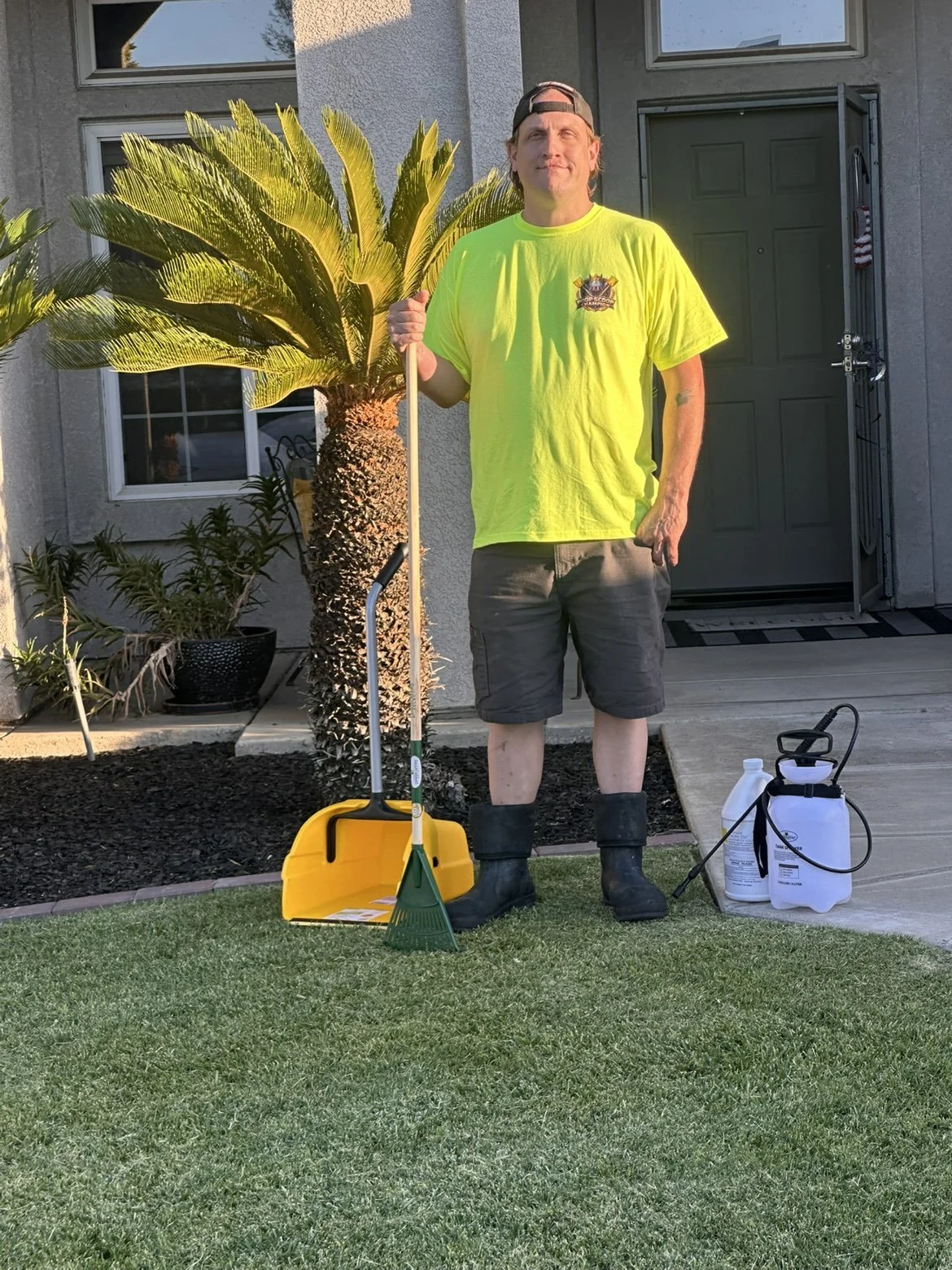 A man in a bright yellow T-shirt and gray shorts stands outside in front of a house, holding a pooper scooper, with a rake and kennel grade disinfectant . He is wearing black rubber boots and a backward cap, standing on a grassy lawn.