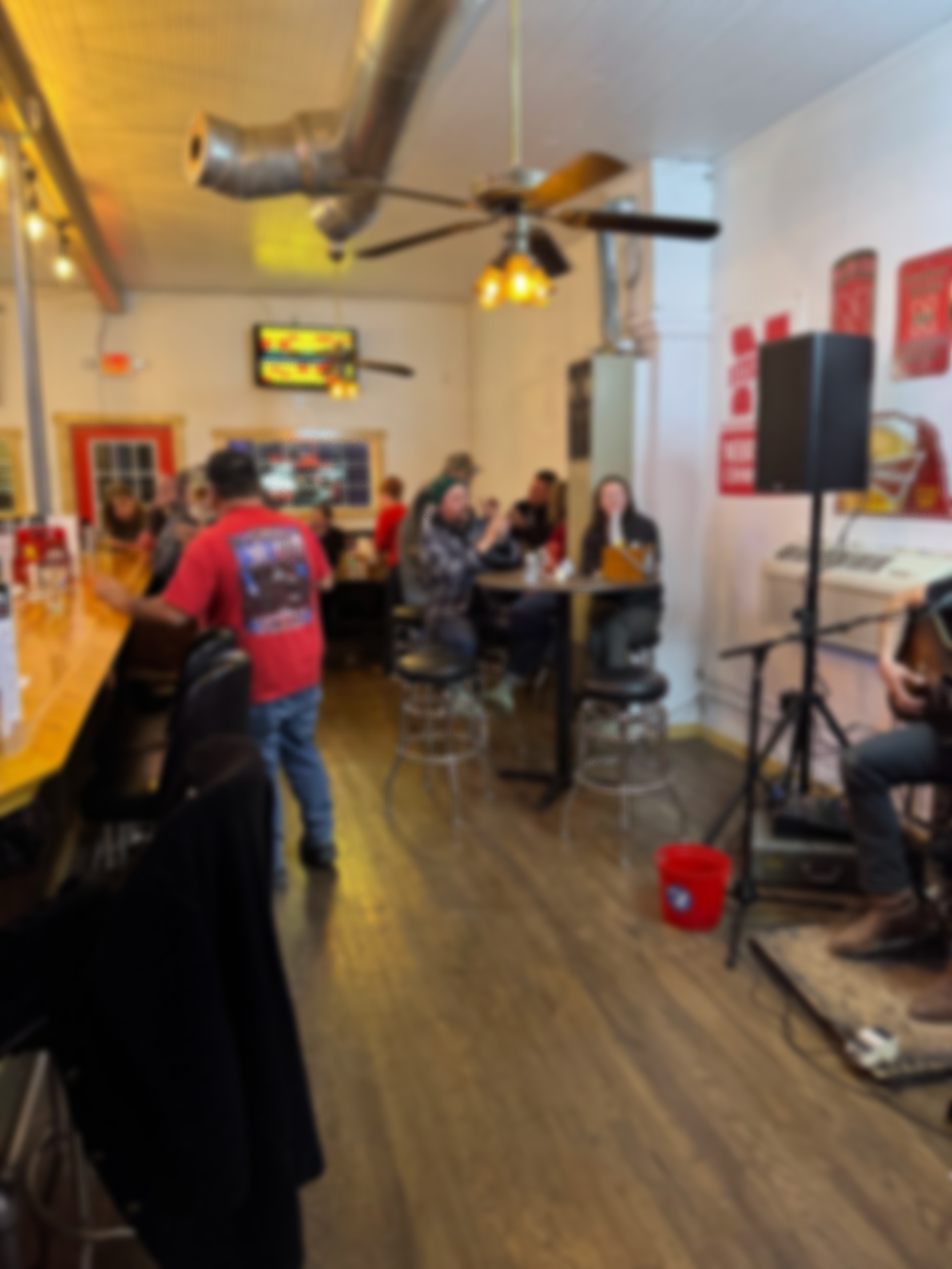 A crowded restaurant with people sitting at tables, a ceiling fan, and a television on the wall. The floor is wooden, and there is a speaker and cleaning bucket in the corner.