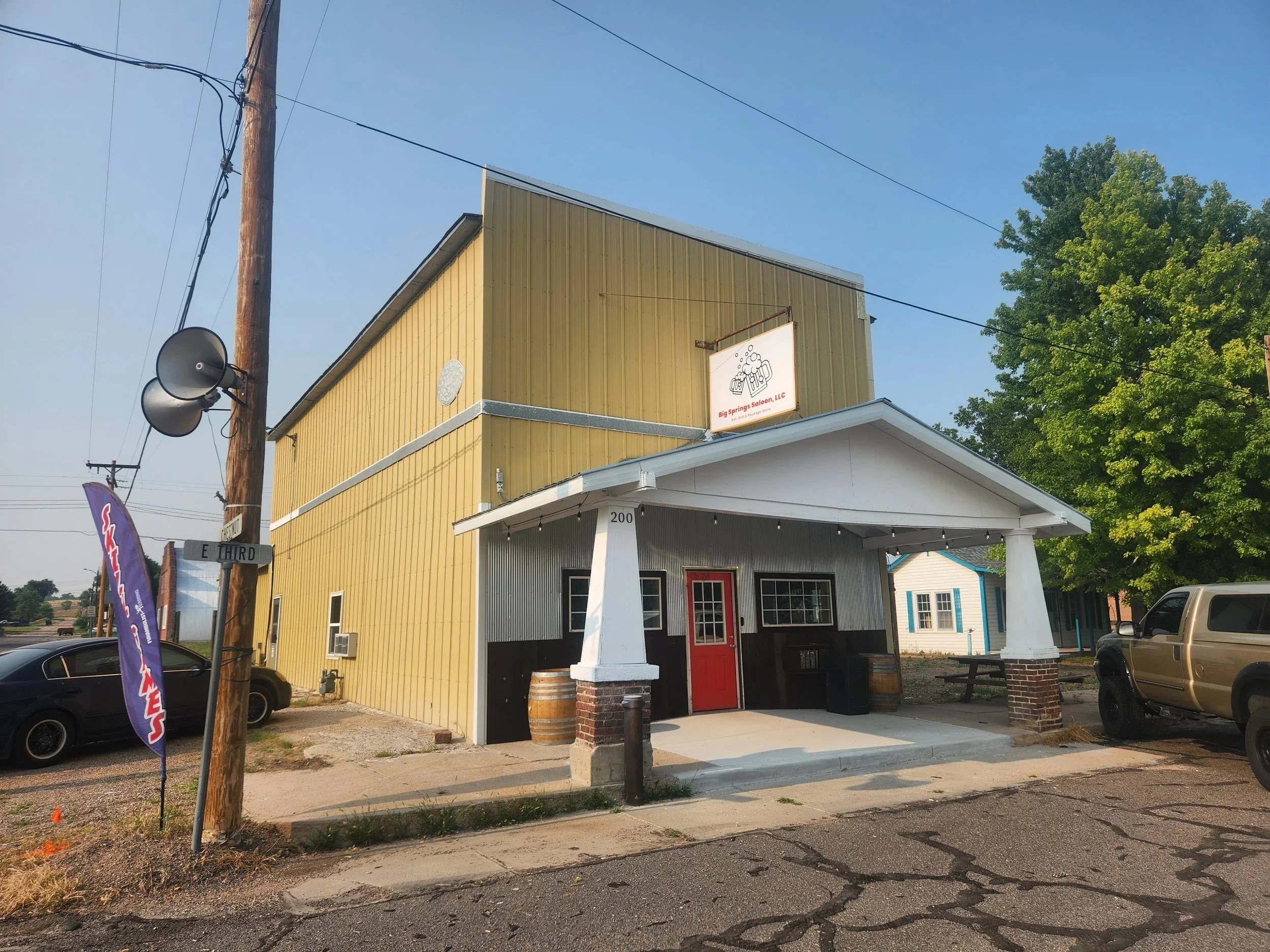 Yellow restaurant building with a red door, white trim, and a sign that reads 'Big Springs Saloon, LLC' with beer mugs. Outdoor seating includes a bench and table, with some barrels and plants nearby. Overhead string lights are visible, with a large green tree to the right and a utility pole with attached speakers on the left.