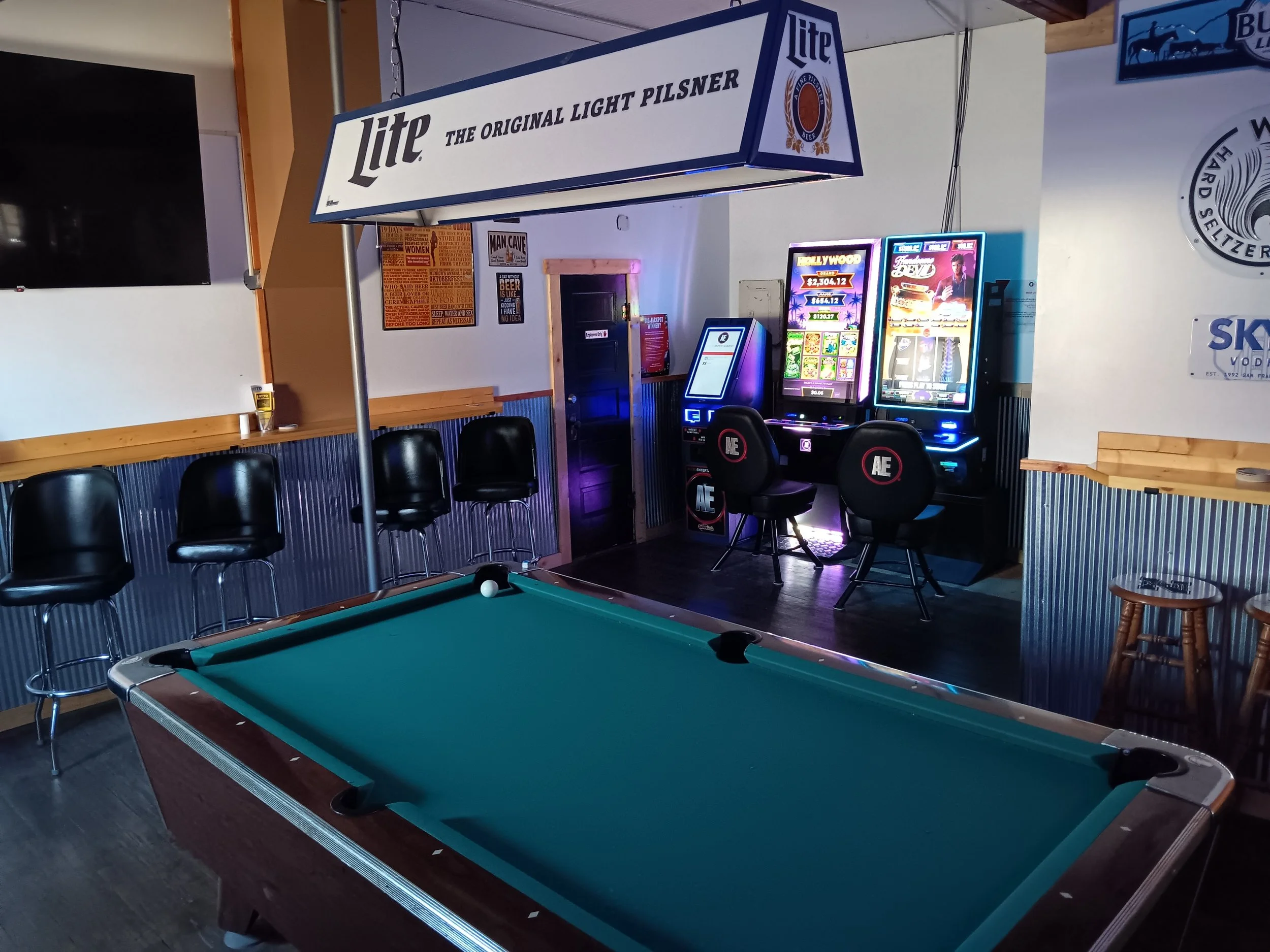 Indoor bar or game room featuring a pool table in the foreground, electronic gaming machines, black chairs, and a large Miller beer sign hanging from the ceiling.