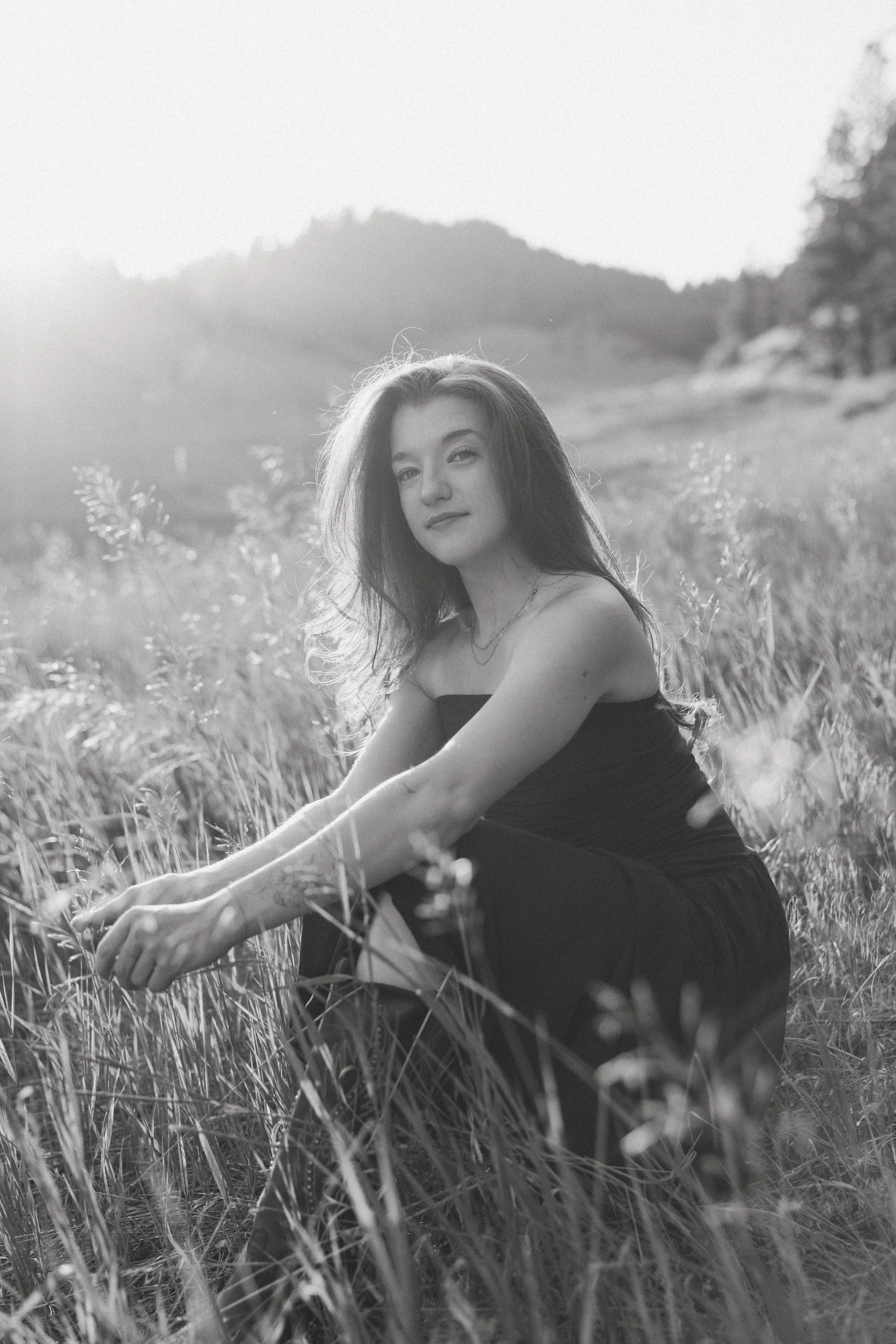 Black and white photograph of a young woman sitting in a field of tall grass with mountains and trees in the background, looking at the camera.