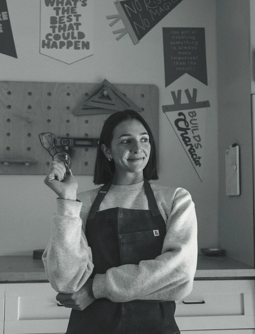 A woman wearing a dark apron and a light sweatshirt, holding glasses, standing in a workspace with motivational posters on the wall.