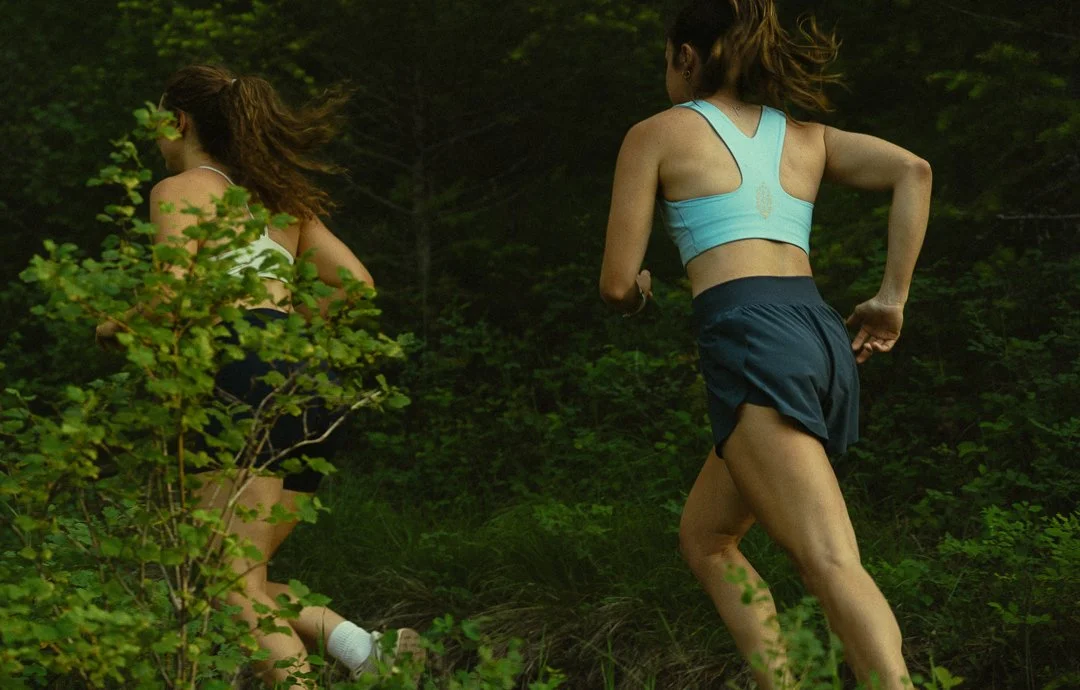 Two women running outdoors in a wooded area, wearing workout clothes.