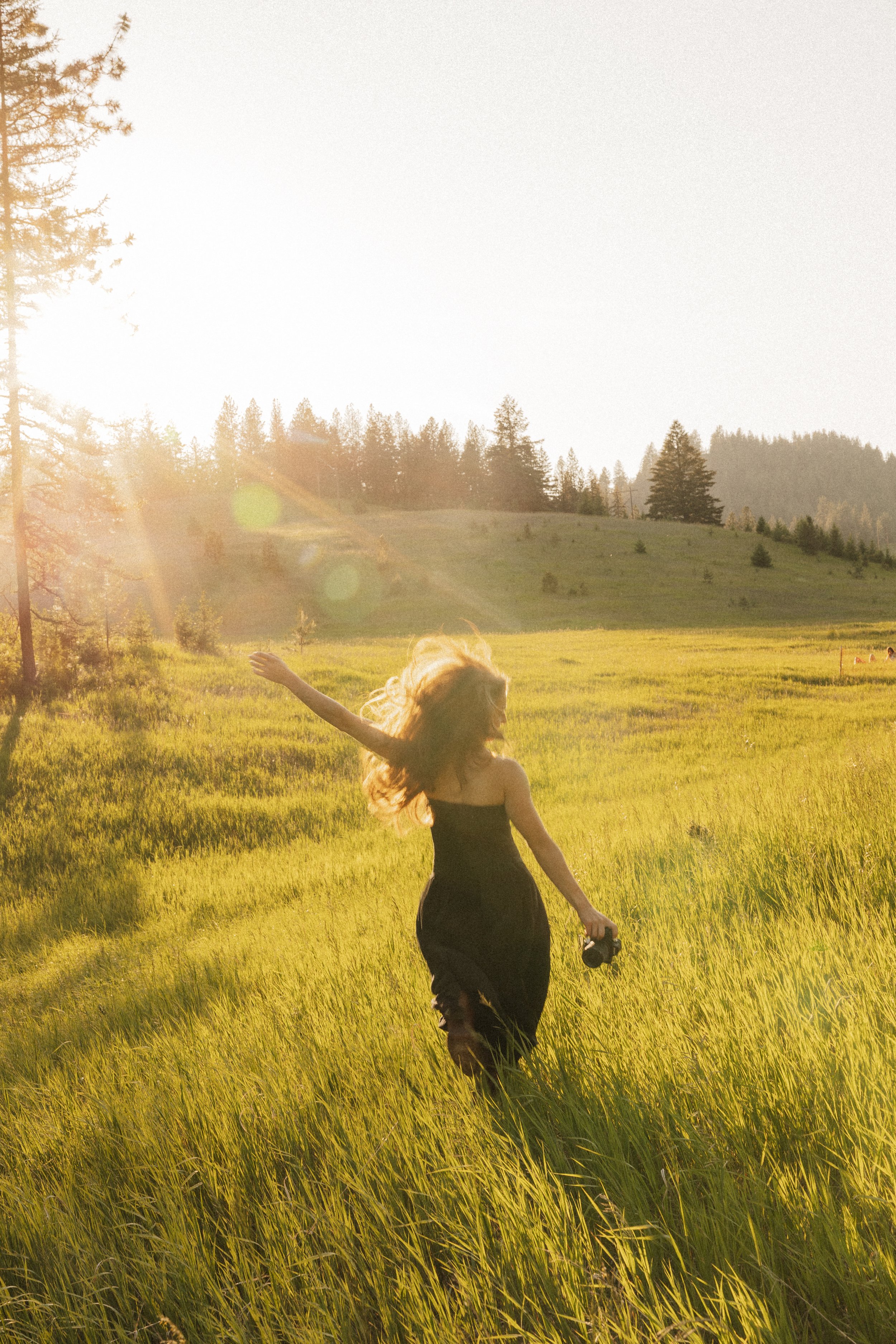 A girl with long hair in a black dress running through a green field during sunset, holding sunglasses in her left hand, with a backdrop of trees and hills.