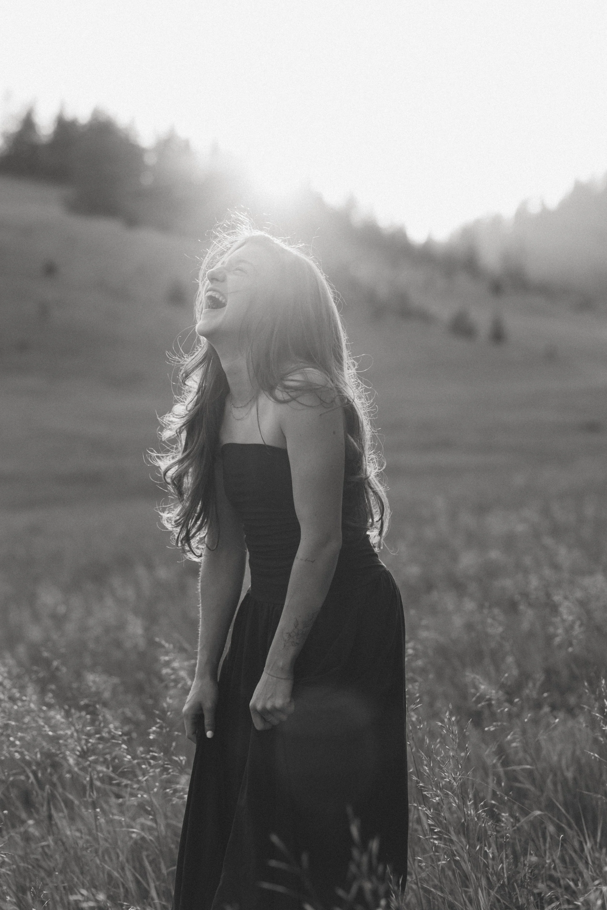 A woman laughing and standing in a field of tall grass with hills and trees in the background, captured in black and white.
