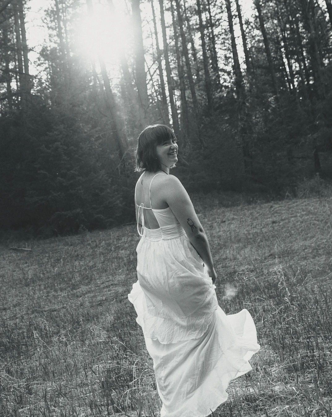 A woman in a white dress walking through a grassy field with a forest in the background, sunlight filtering through the trees, black and white photograph.