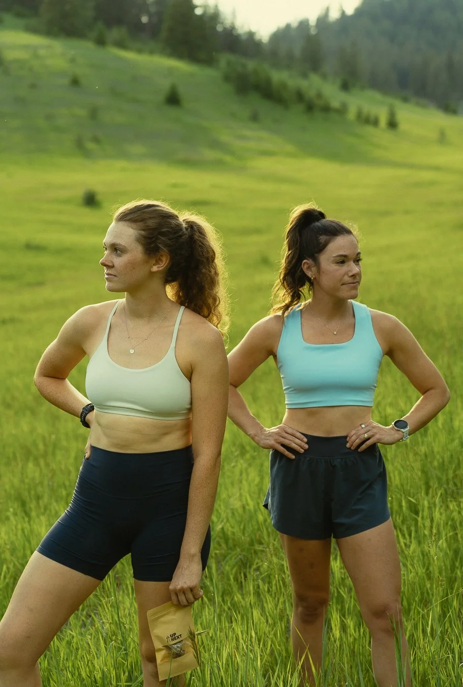 Two women standing in a grassy field with green hills in the background. They are dressed in athletic workout clothes, one in a light green sports bra and black shorts, the other in a blue sports bra and black shorts. They are looking in opposite directions.