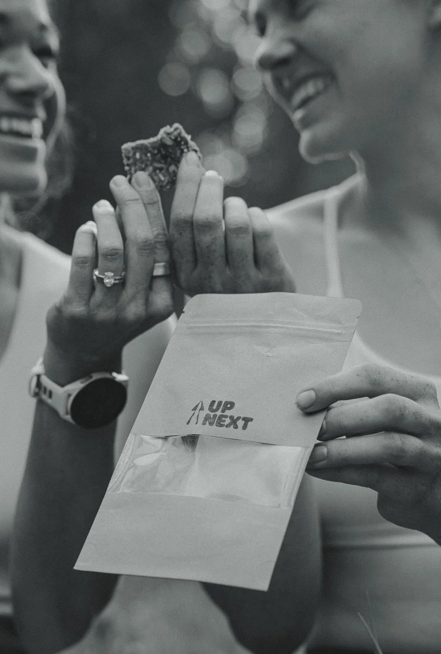 Two women sharing a smile as one offers a piece of food, possibly a cookie, to the other. One woman has a watch and engagement ring, while the other holds a bag or envelope labeled 'UP NEXT' with an arrow pointing upward.