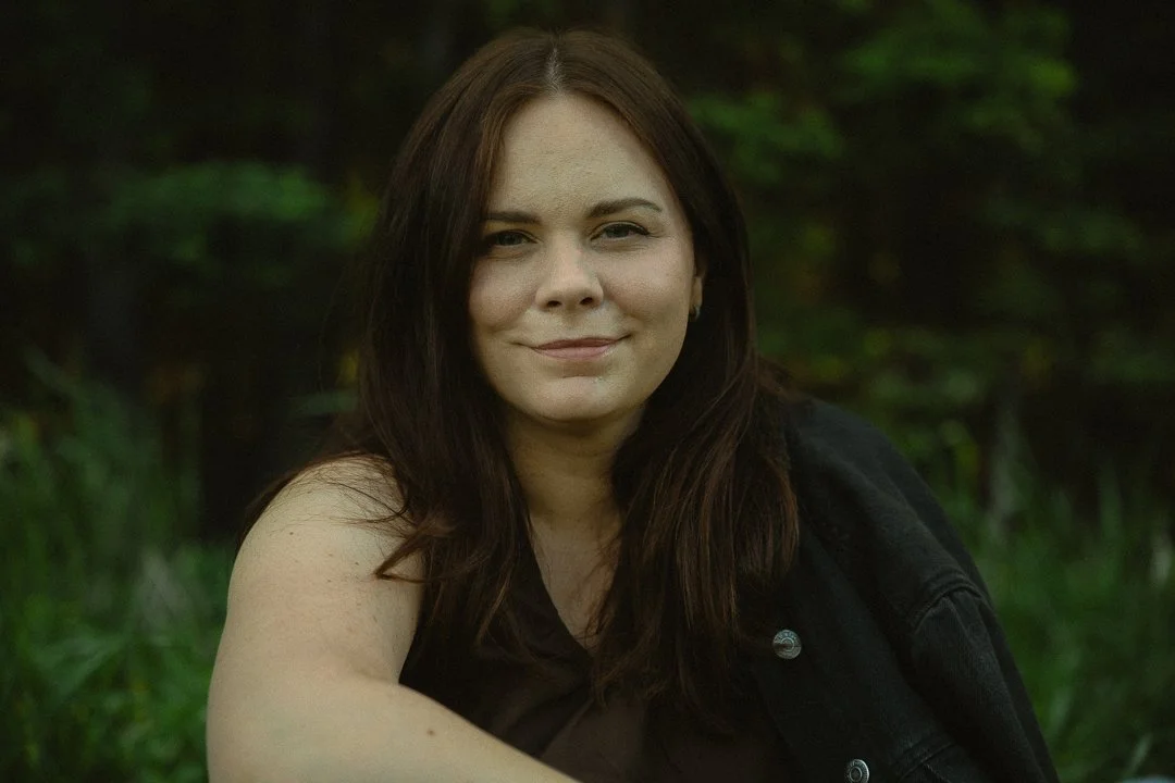 A woman with long dark hair and fair skin sitting outdoors with greenery in the background, wearing a black sleeveless top and a black jacket, smiling softly.