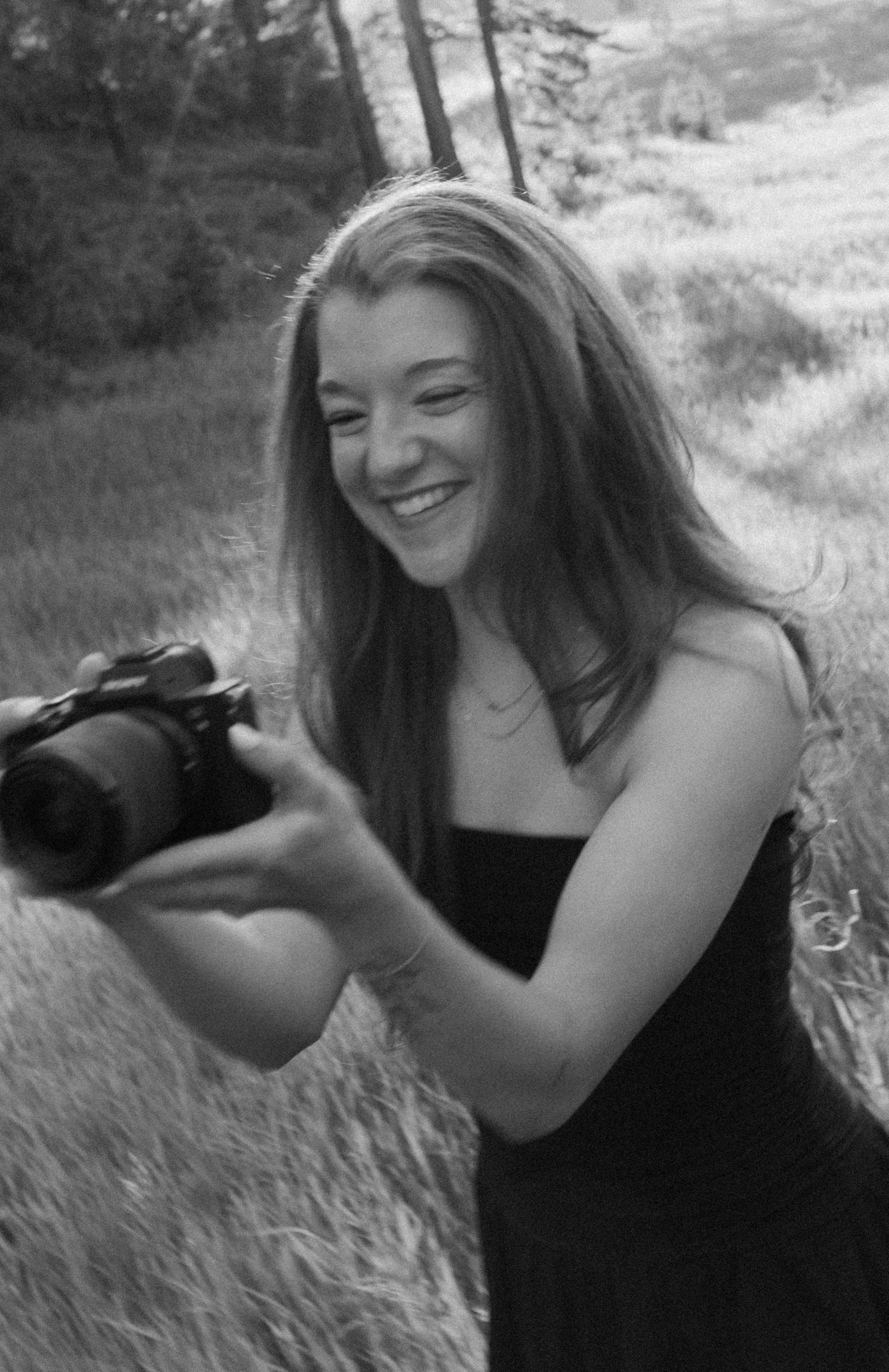 A young woman with long hair smiling and aiming a camera, sitting on a grassy field with trees in the background, in black and white.