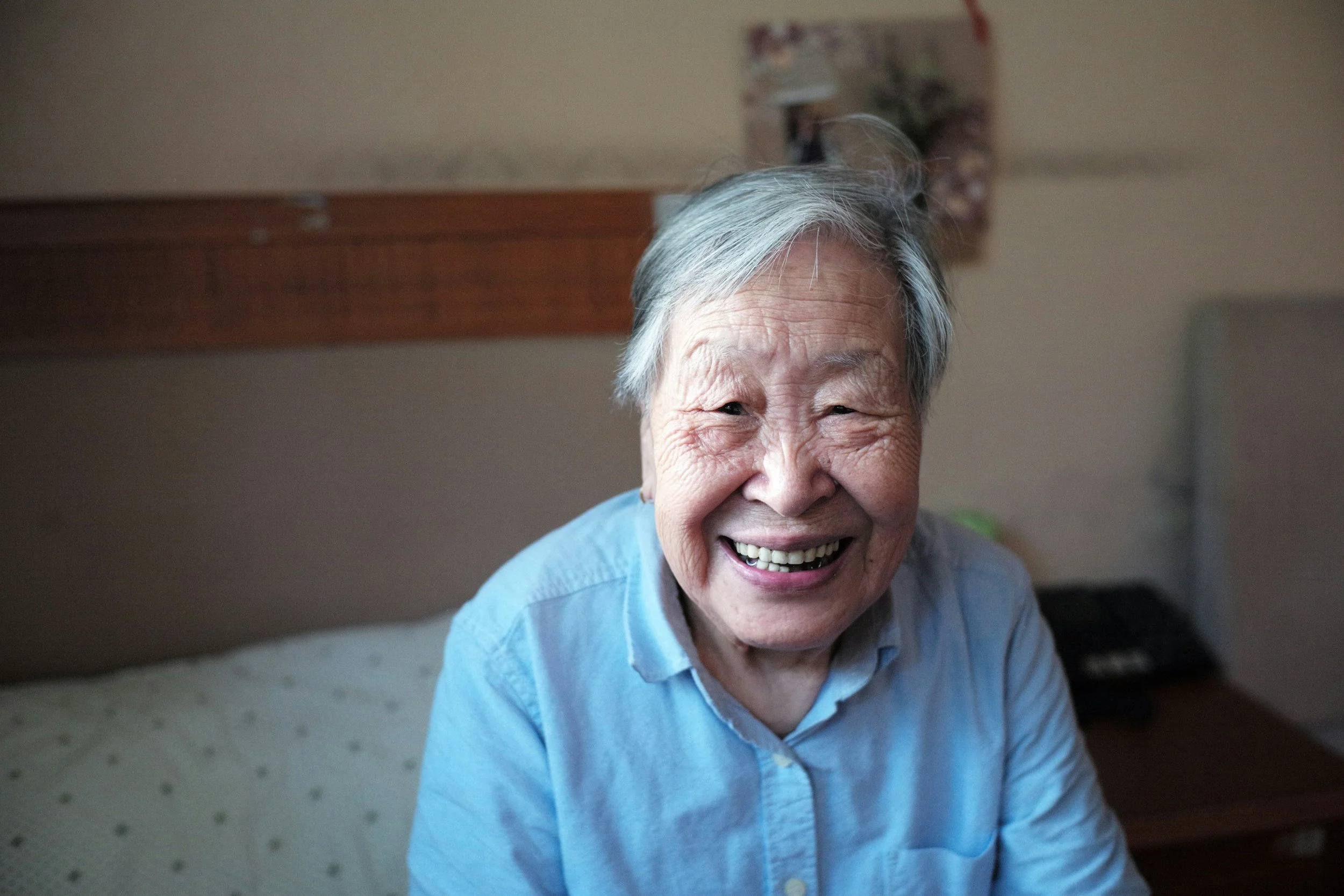 A smiling elderly woman with gray hair, wearing a light blue shirt, sitting indoors in a cozy room.