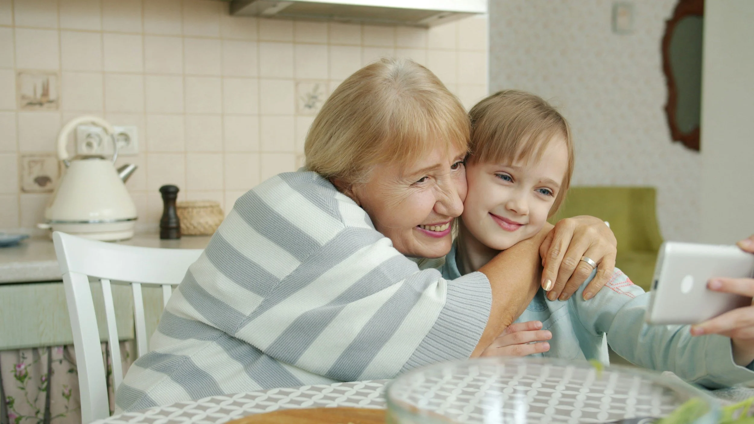 A grandmother and a young boy taking a selfie together at a dining table in a kitchen, both smiling.