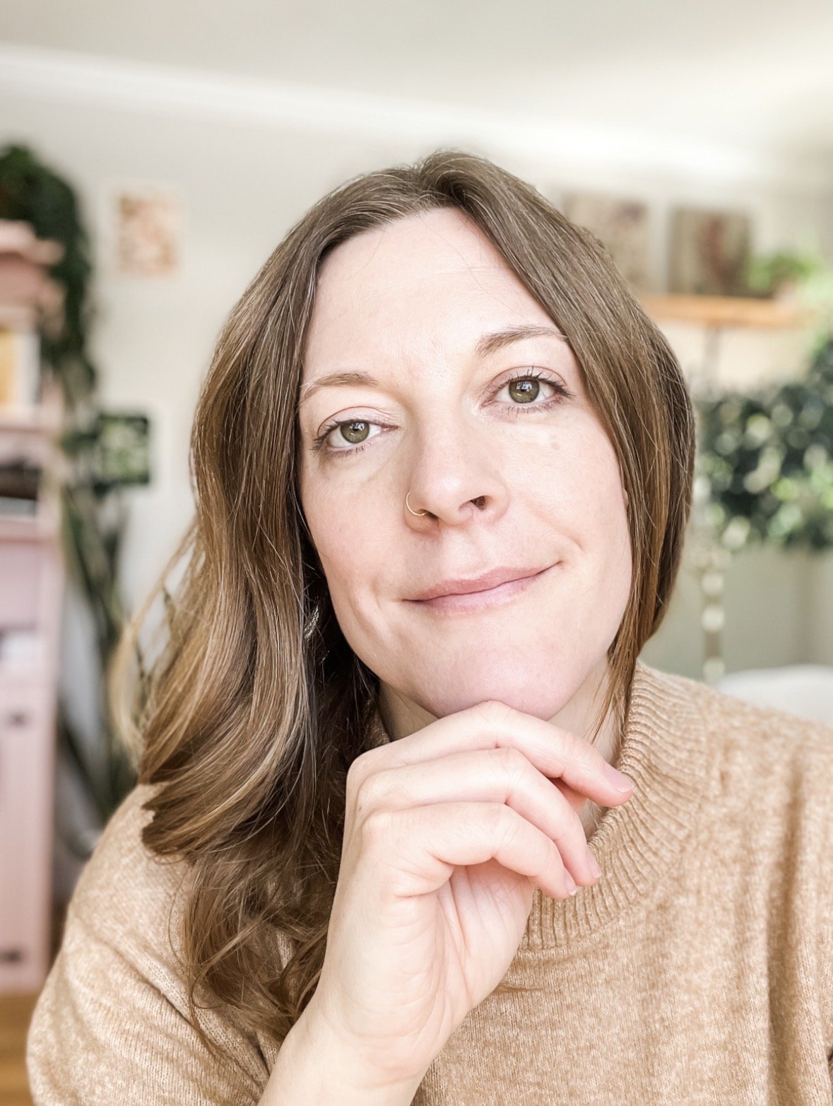A woman with shoulder-length brown hair, green eyes, and a nose ring rests her chin on her hand inside a well-lit room with green plants and shelves in the background.