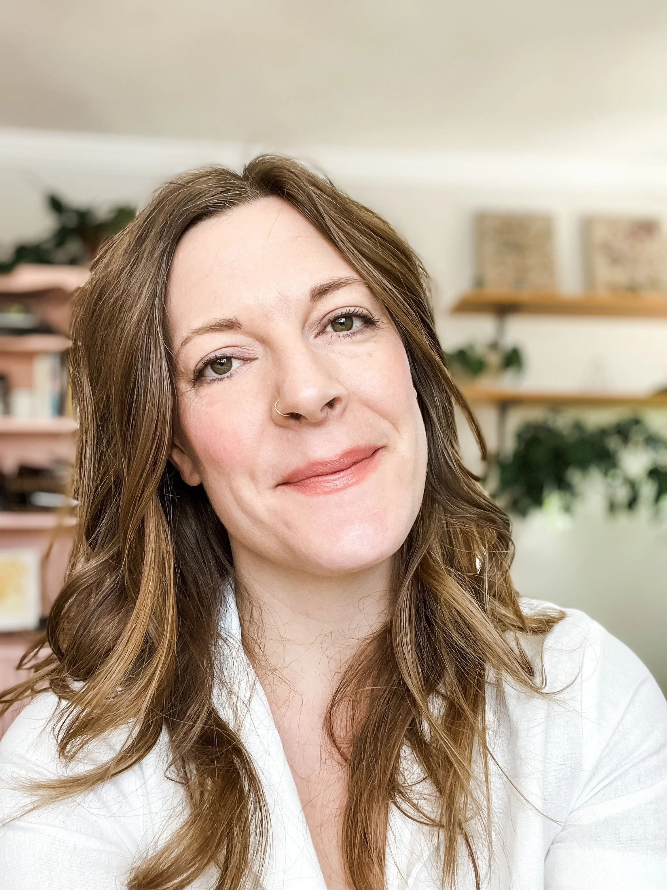 A woman with long, wavy brown hair and light skin smiling softly, wearing a white shirt, in a room with shelves and plants in the background.