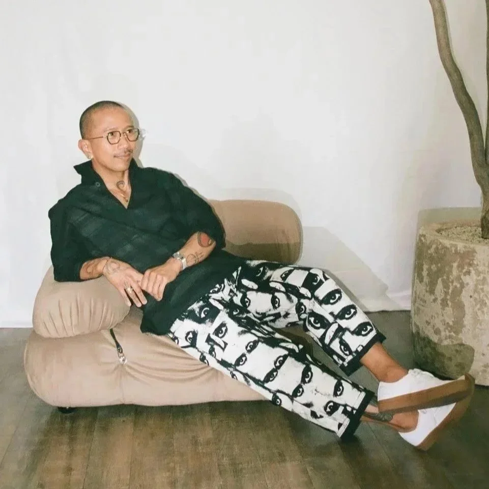 A person with short hair, glasses, and multiple tattoos sitting on a beige cushion sofa, wearing black top, patterned pants with eye designs, and white platform shoes, next to a tall potted plant in a minimalist room.