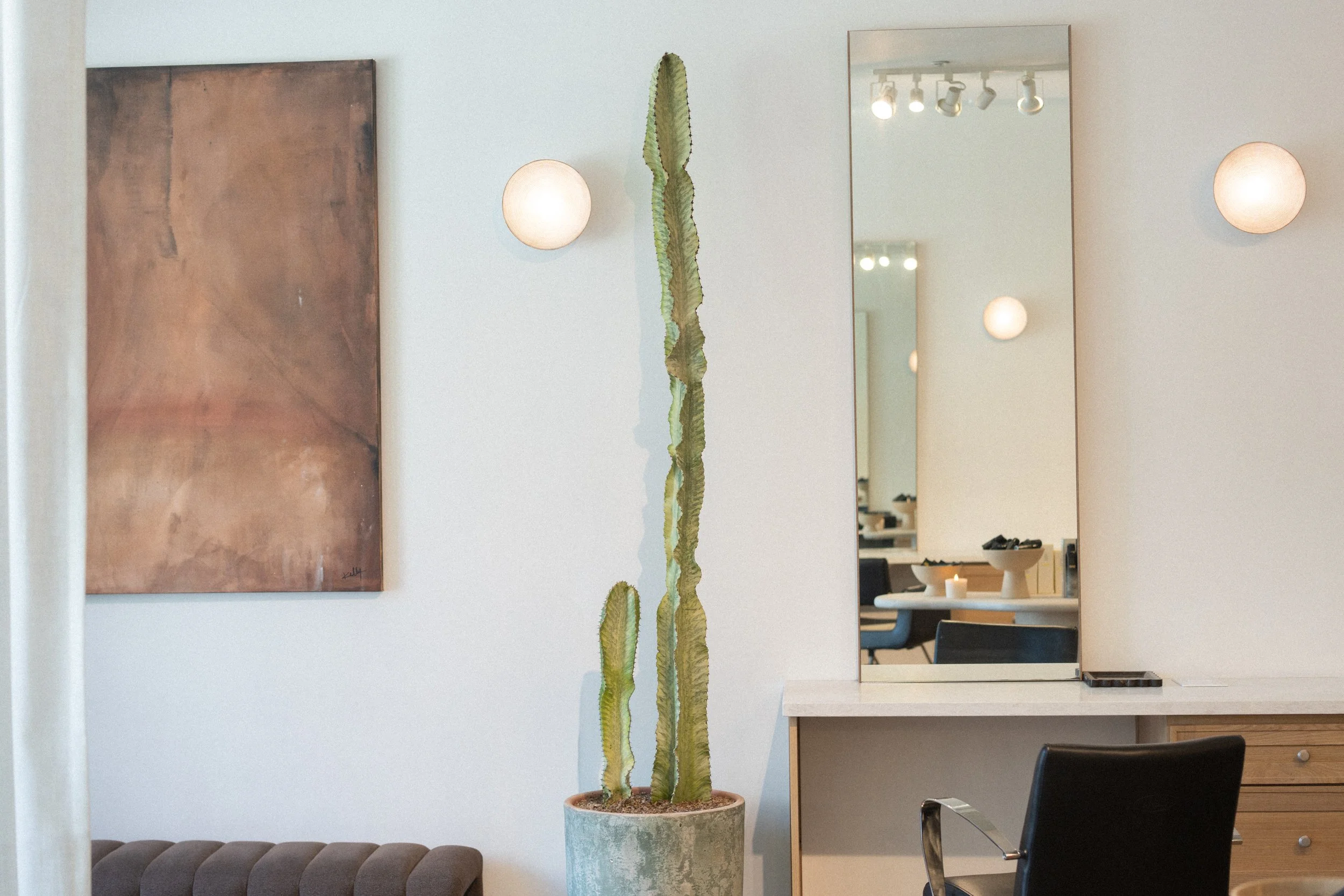 Salon styling station with a tall mirror, glowing globe sconces, a tall cactus plant, and abstract wall art above a light wood cabinet.