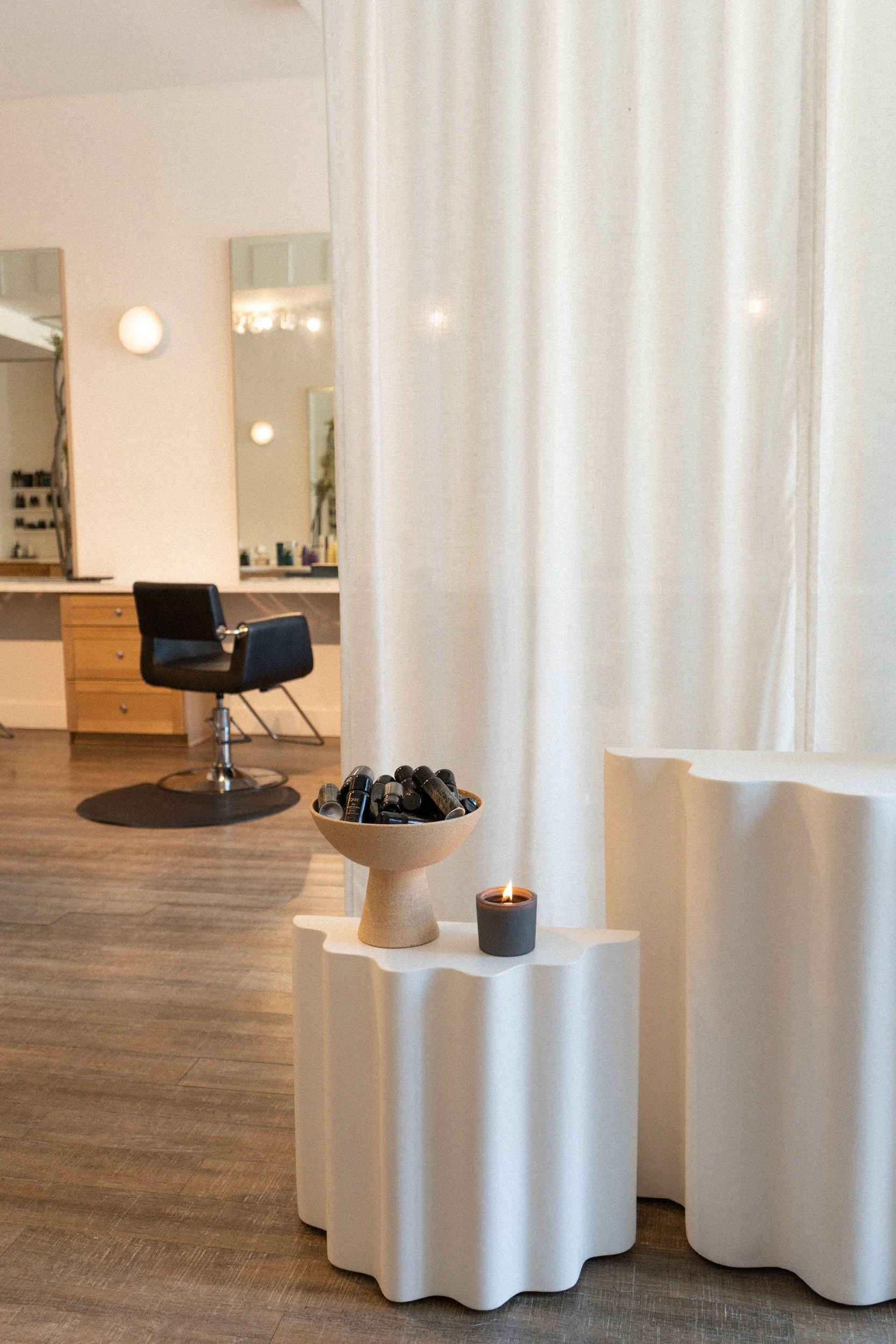 Cozy salon corner with sheer white curtains, a small draped pedestal table holding a decorative bowl and candle, and a black styling chair glowing softly in the background.