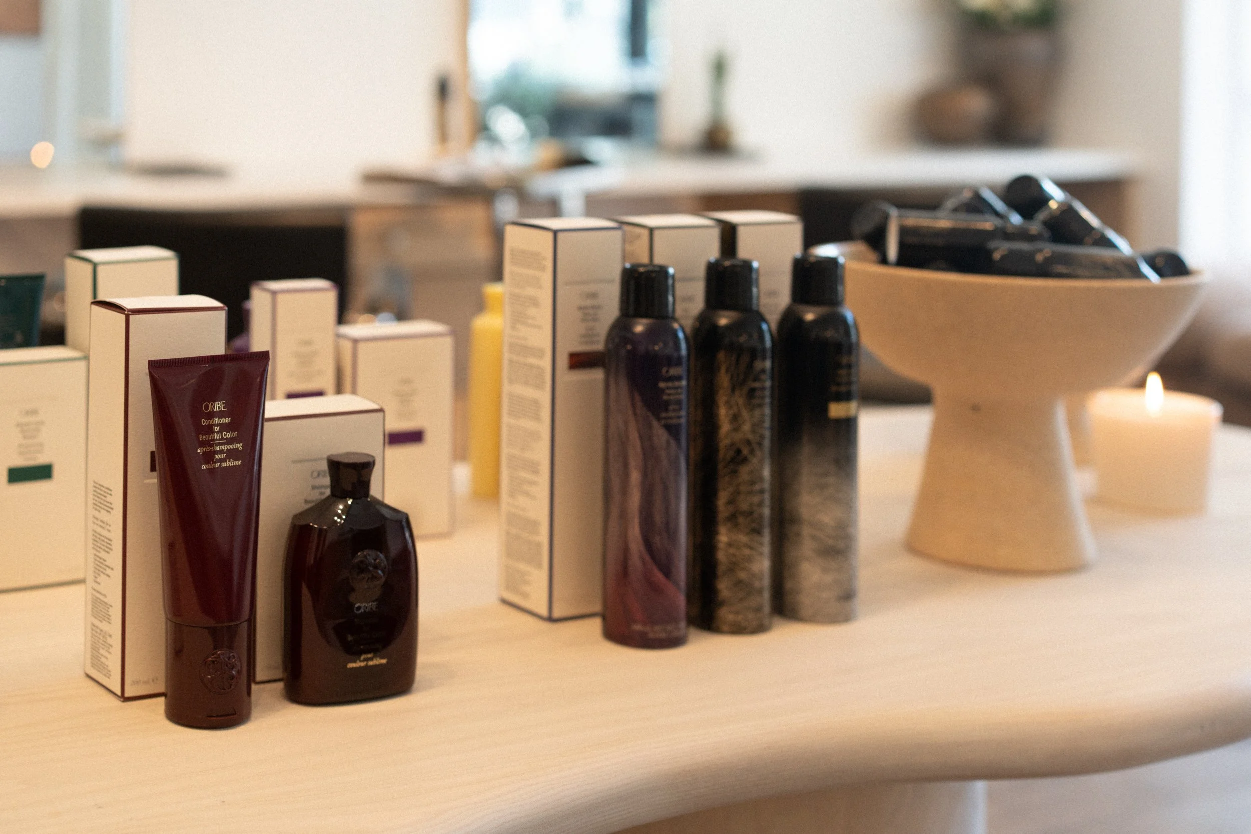 Oribe retail display with dark shampoo and styling bottles, product boxes, and a lit candle arranged on a salon counter.