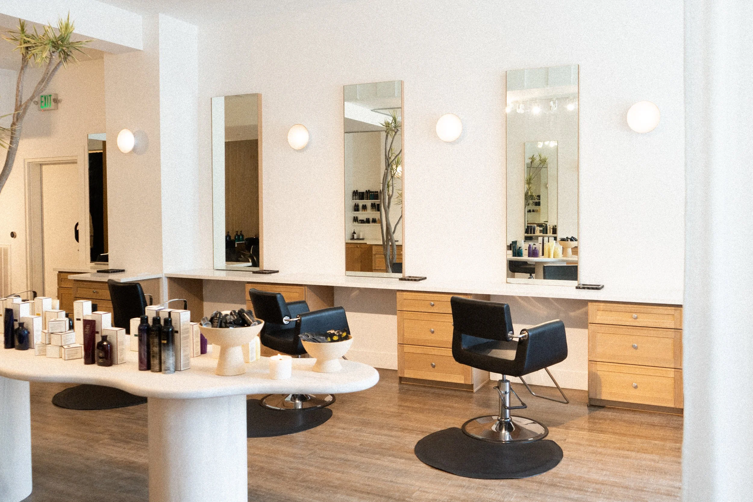 Bright, modern salon interior with black styling chairs at tall mirrors, light wood stations, and a curved white counter displaying hair products from Oribe and K18 in the foreground.