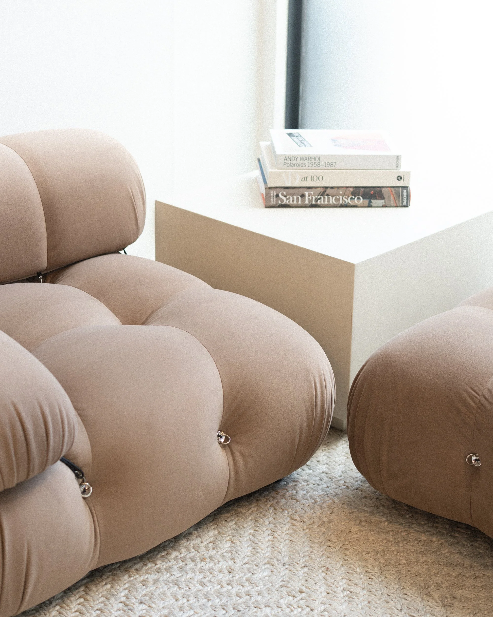 Salon waiting area with a plush beige tufted lounge chair beside a small side table stacked with books, in soft natural light.