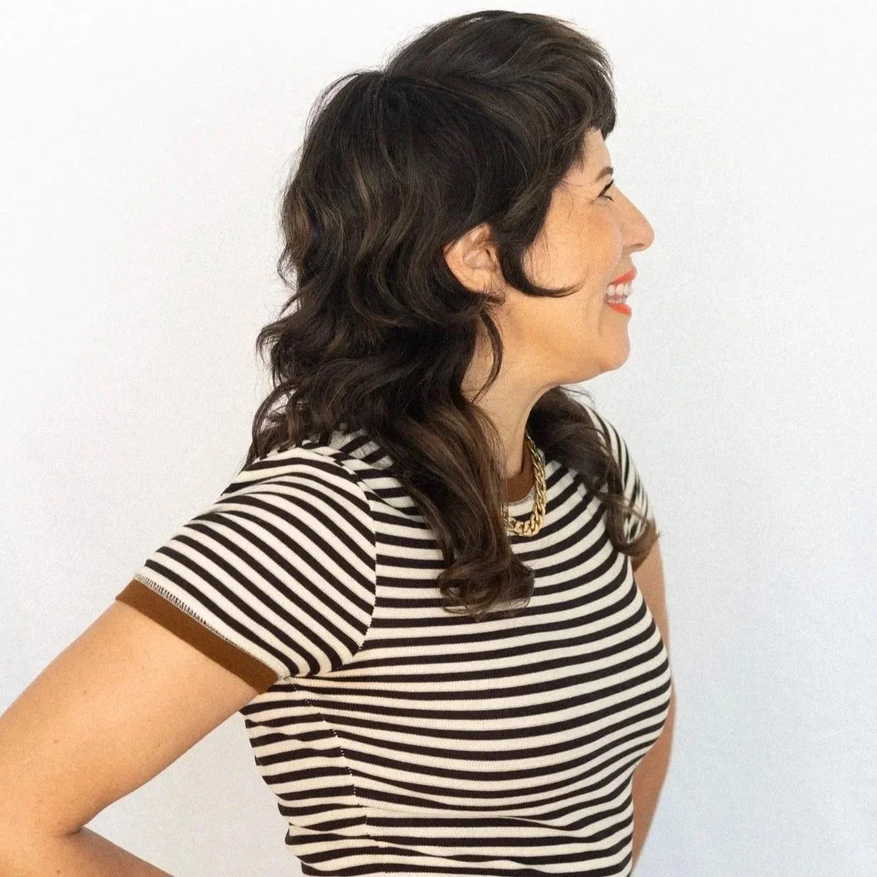 A woman with rich brunette shag haircut, wavy hair, smiling and wearing a black and white striped shirt, standing against a plain white background.