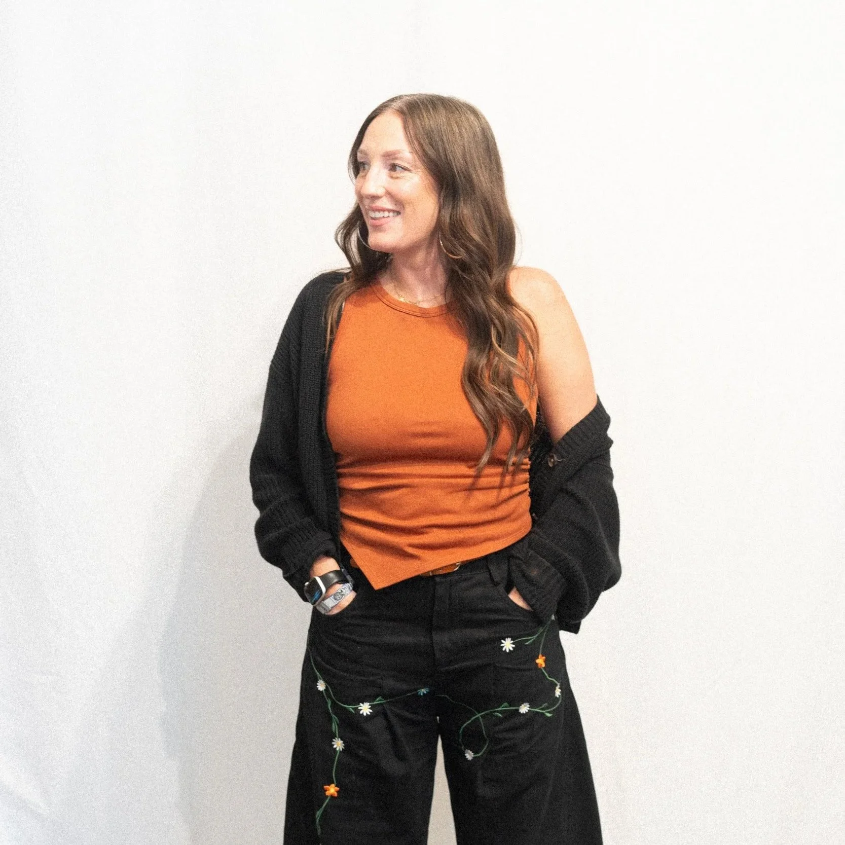 A woman with long wavy brown hair smiling and looking to the left, wearing a sleeveless top and a black cardigan, standing against a plain white wall.