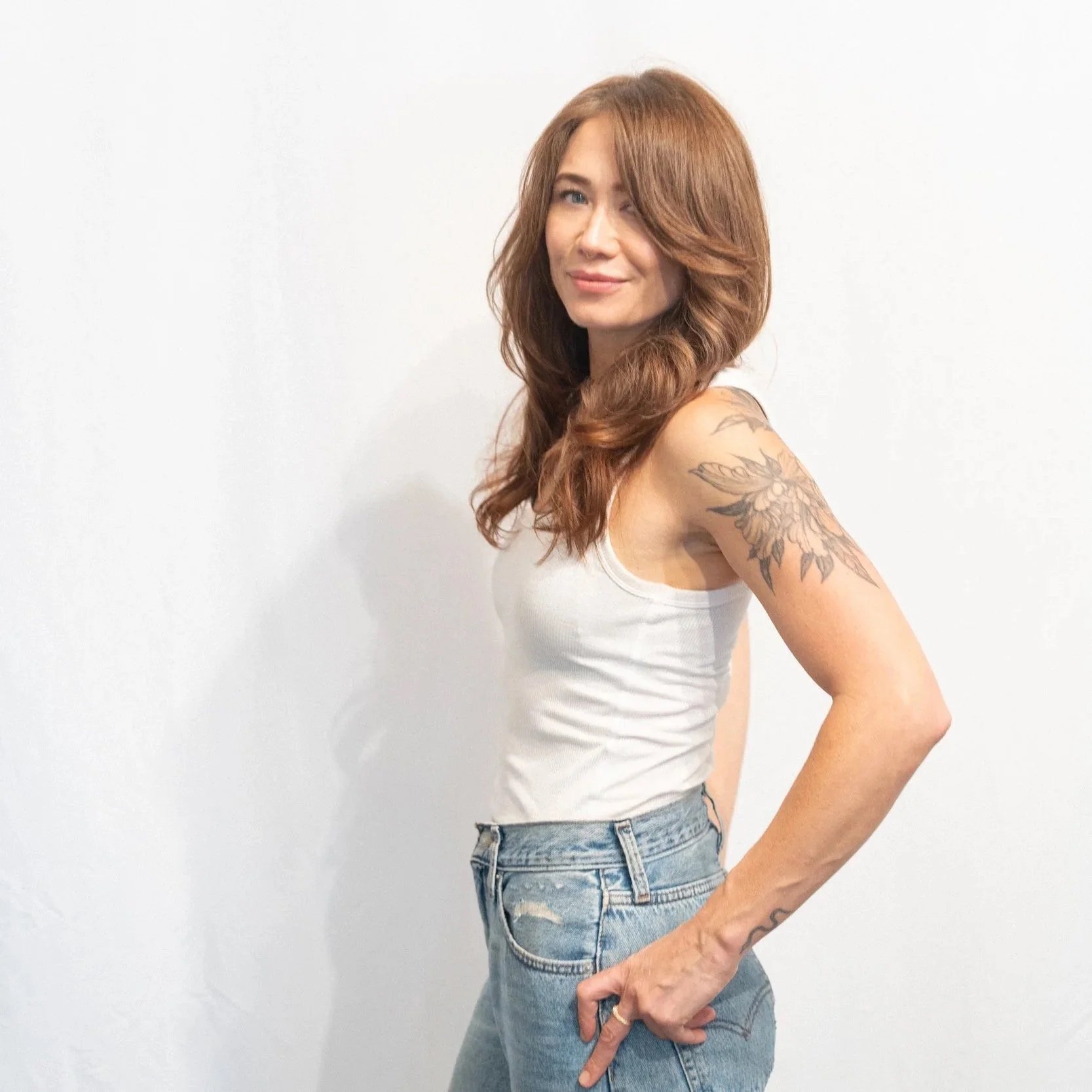 A young woman with copper hair, wearing a white tank top and blue jeans, showing her arm tattoo and holding her waist, standing against a plain white wall.
