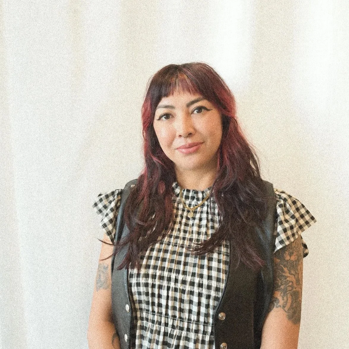 A woman with dark red hair and tattoos on her arms, wearing a black leather vest over a black and white checkered shirt, standing against a plain white background.