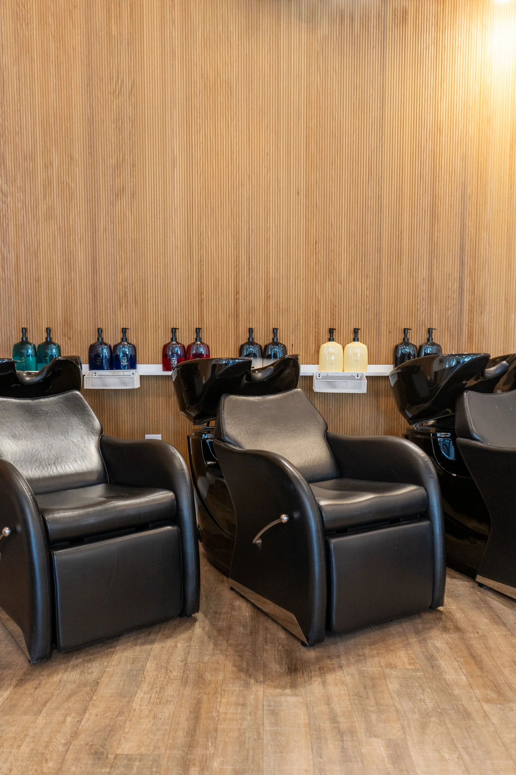 Salon shampoo area with black reclining shampoo chairs against a wood-paneled wall, shelves above stocked with Oribe shampoo conditioner and hair products.