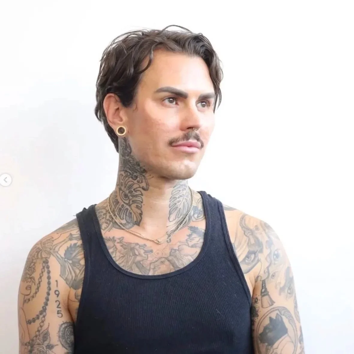 Portrait of a tattooed man with dark hair, wearing a black tank top, earrings, and necklaces, standing against a light background.