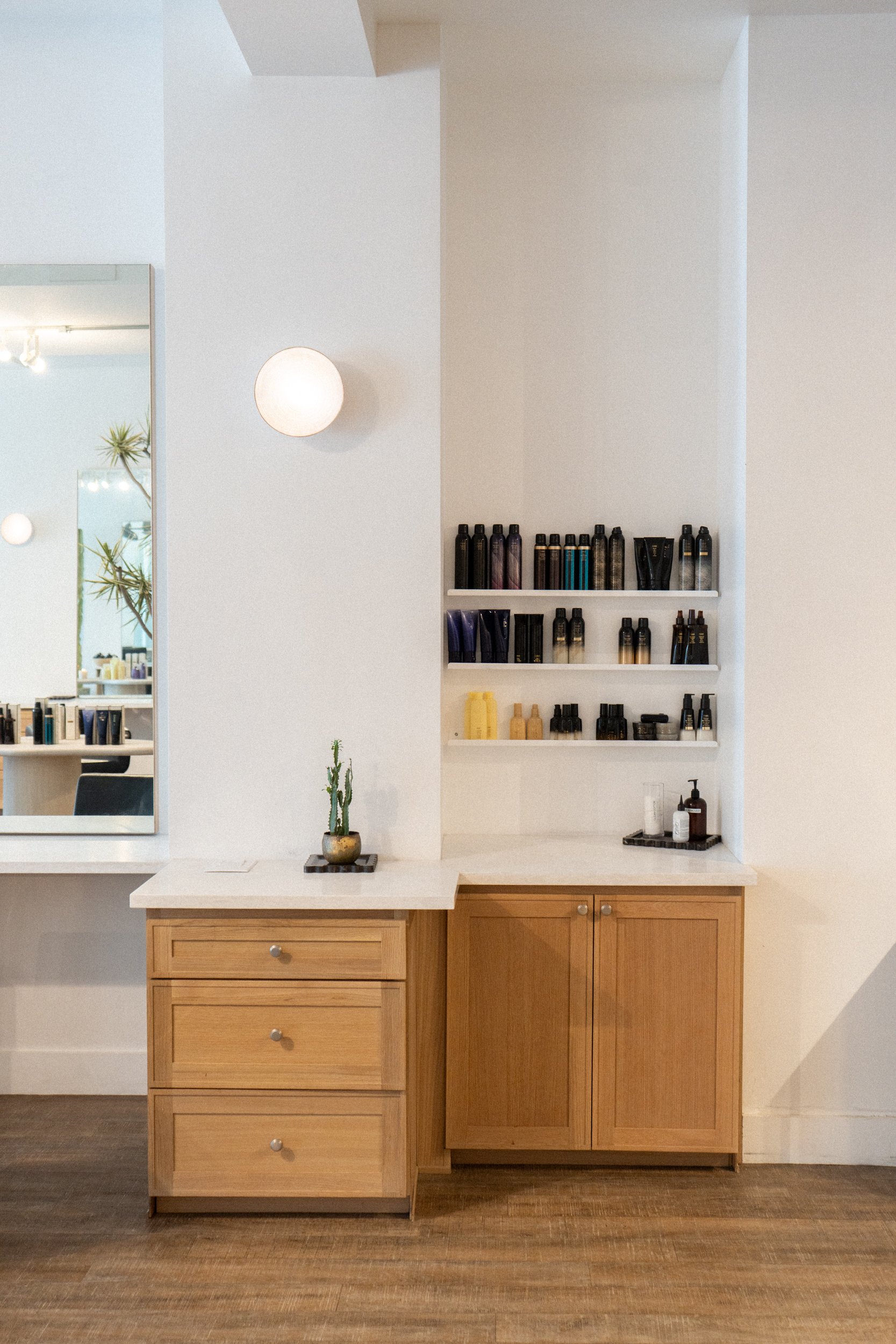 Salon back bar with oribe and k18 hair products light wood cabinets, open shelves stocked with hair products, a mirror, and a warm globe light glowing above on dark wood floors.