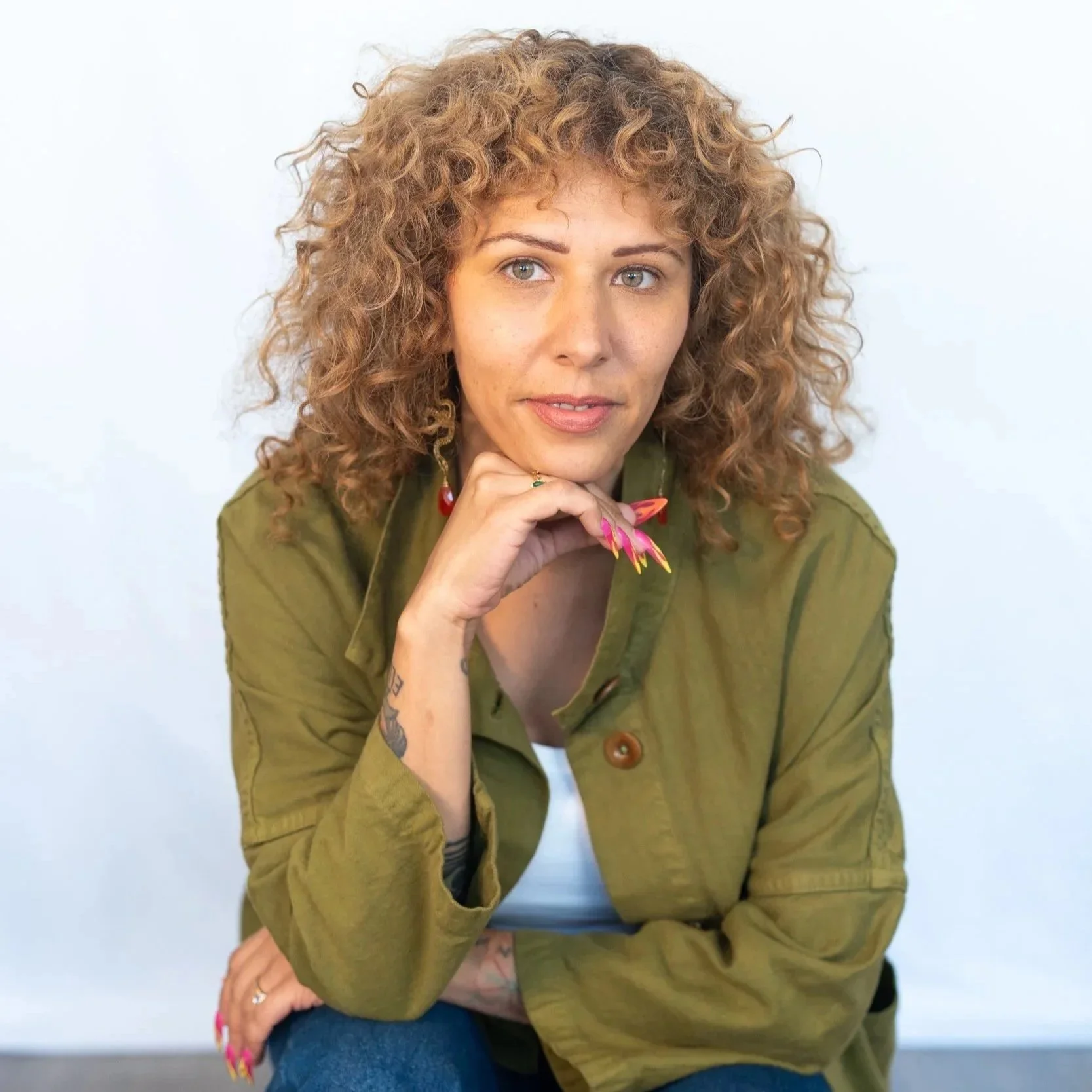 A woman with curly hair, wearing a green jacket over a white top, sits with her chin resting on her hand, looking into the camera.