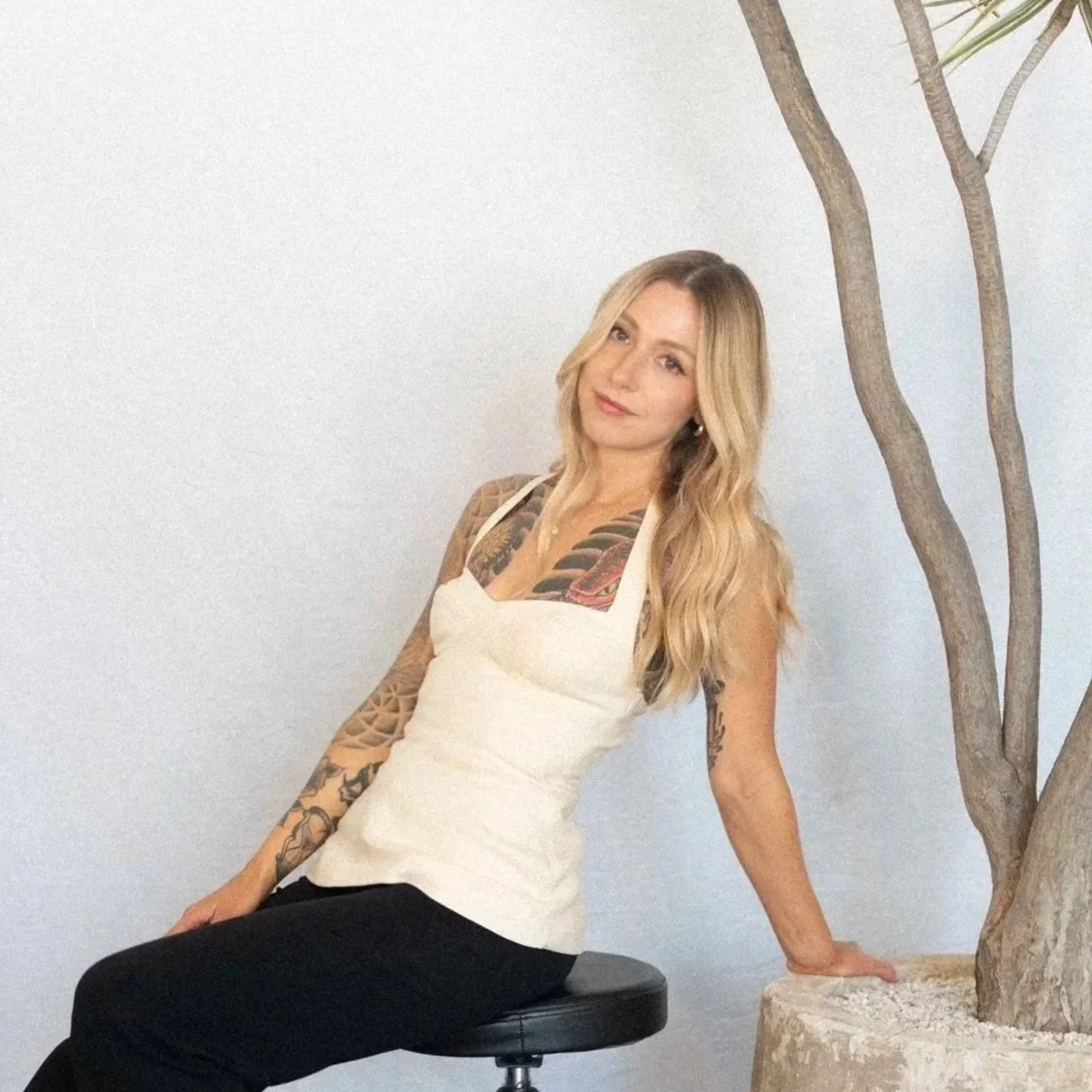 A woman with long golden blonde hair, tattoos on her arms and chest, wearing a white sleeveless top and black pants, sitting on a black stool next to a potted tree with a white wall background.