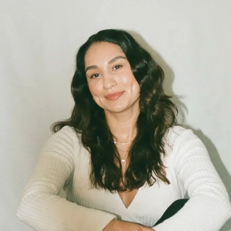 A woman with long, wavy dark espresso hair smiling at the camera, wearing a white ribbed sweater and layered necklaces, sitting against a plain wall.