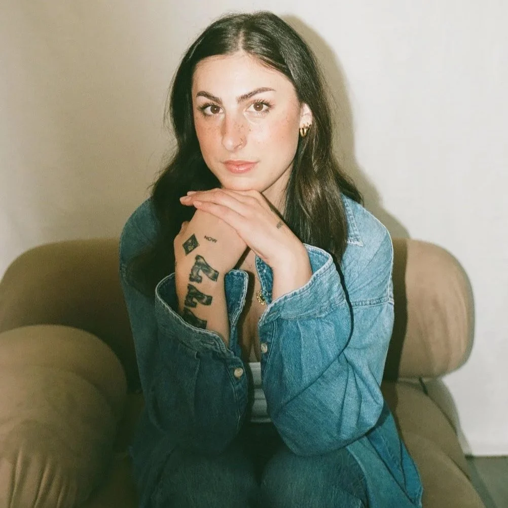 A young woman with dark hair and light skin, wearing a denim jacket, sitting on a beige couch, with her hands resting under her chin, looking at the camera.
