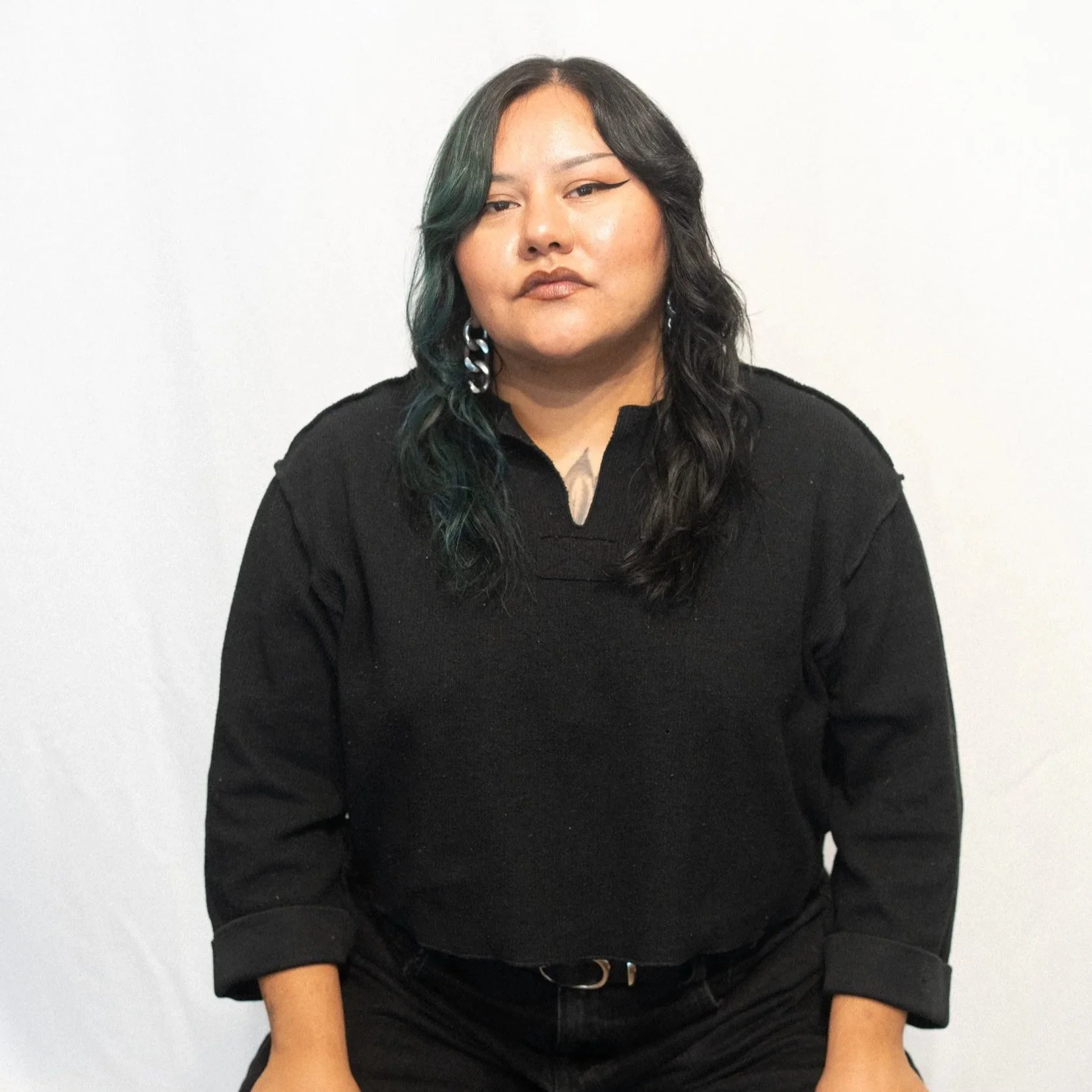 portrait of a woman with shoulder-length dark hair with green highlights, wearing a black long-sleeve top with a small V-neck, silver earrings with a linked chain design, against a plain white wall.