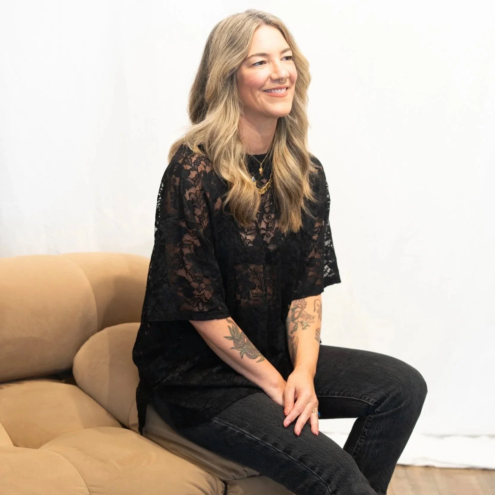 A woman with long wavy blonde hair, wearing a black lace top and black pants, sitting on a beige couch against a plain white wall, smiling and looking to her left.