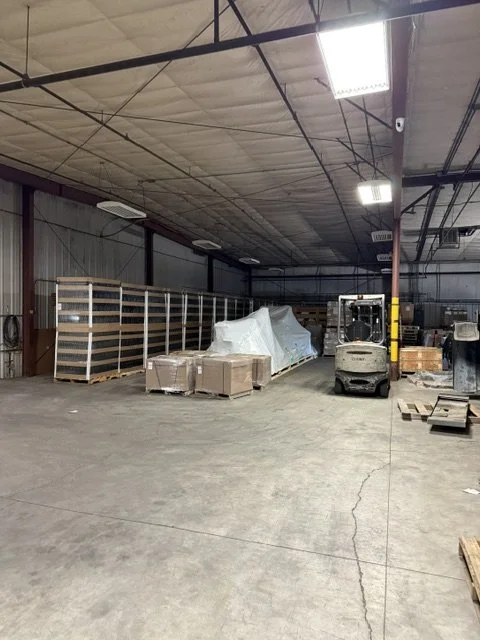 Interior of a warehouse or storage facility with some packing materials, pallets, a forklift, and machinery, with a high ceiling and industrial lighting.