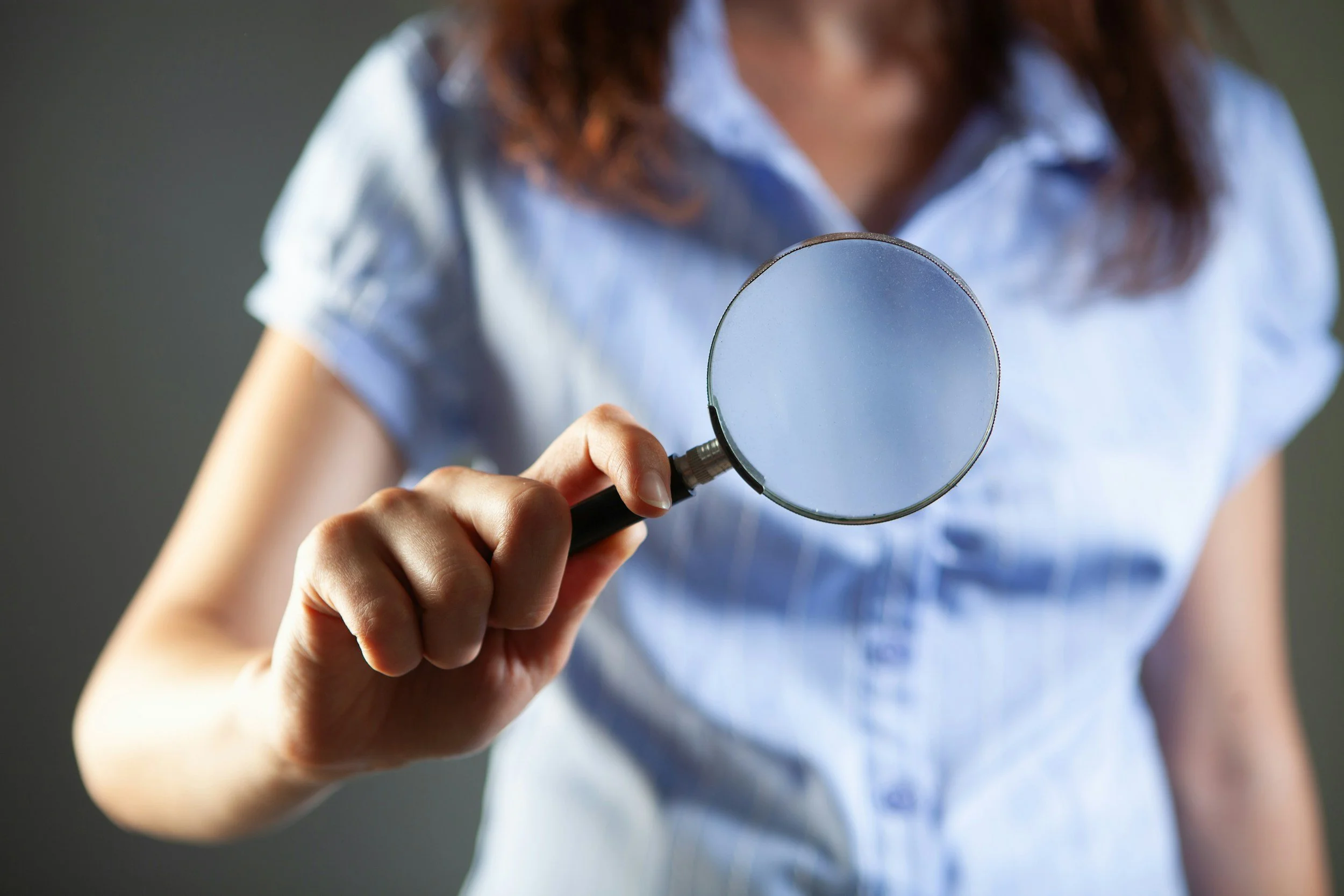 A woman holding a magnifying glass by her chest, wearing a light blue shirt, with reddish-brown hair, against a dark blurred background.