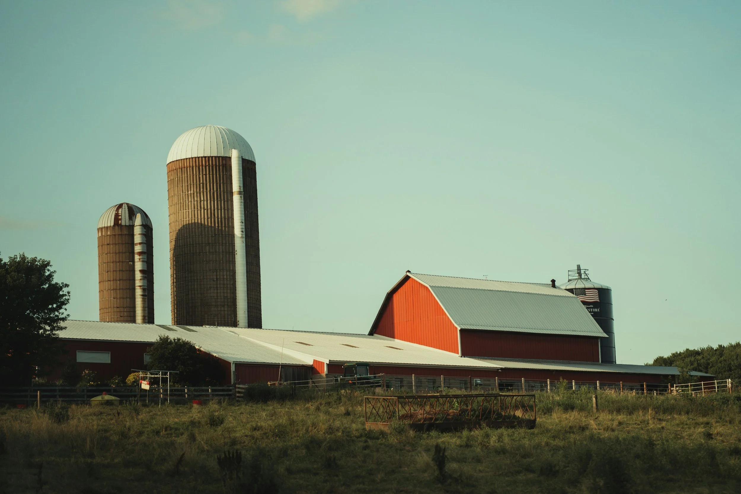 A rural farm scene with two large silos, a red barn, and a half-circle hay storage structure with a metal roof, surrounded by a fenced field and trees in the background under a clear sky.