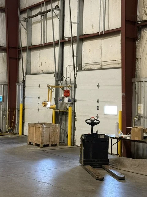 Industrial warehouse with a loading dock, pallet jack, and safety equipment near a large garage door.