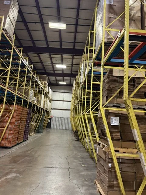 Interior view of a warehouse with tall yellow metal shelving units filled with boxes and items, a concrete floor, and a high ceiling.
