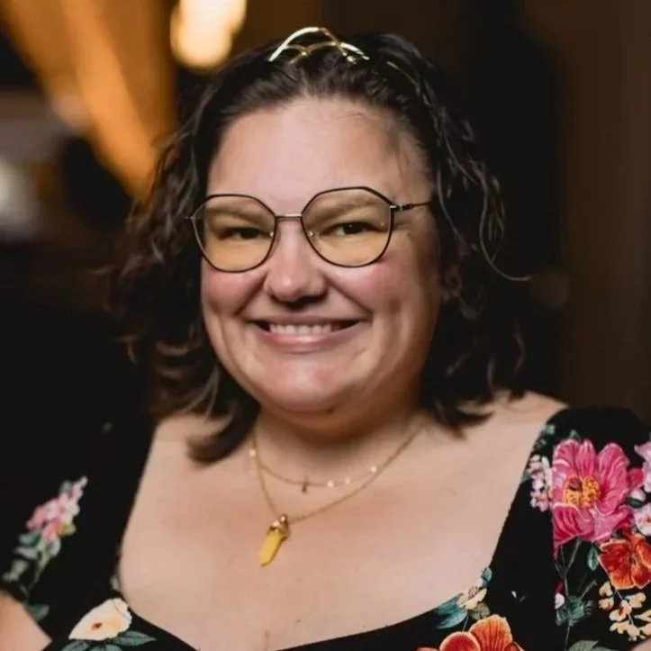 A woman with curly dark hair wearing glasses, smiling, dressed in a floral top, with layered necklaces including one with a yellow pendant.