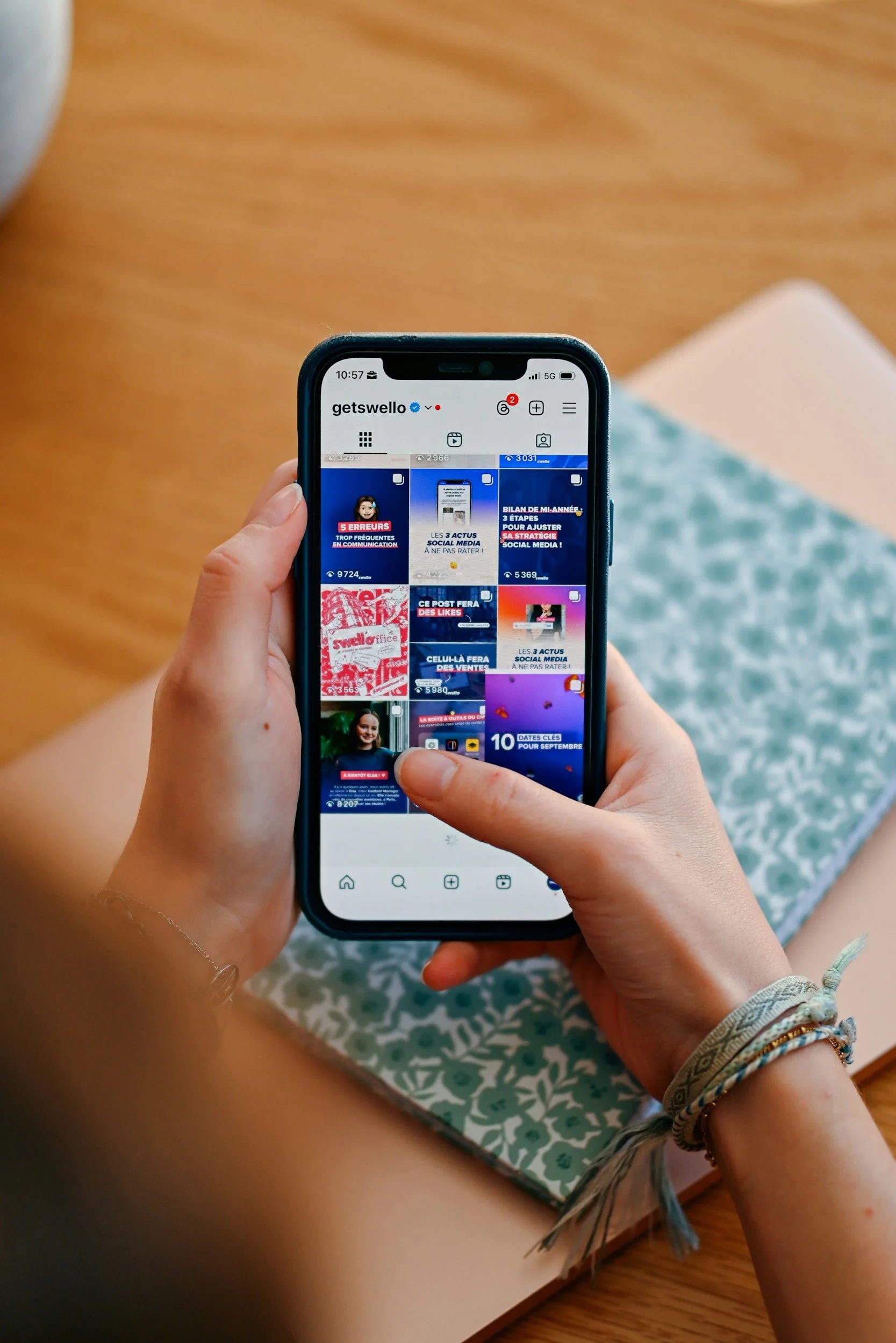 A person holding a smartphone showing an Instagram profile page called 'getswello' with various social media posts and icons, sitting on a wooden surface with patterned fabric and wearing multiple bracelets on the wrist.