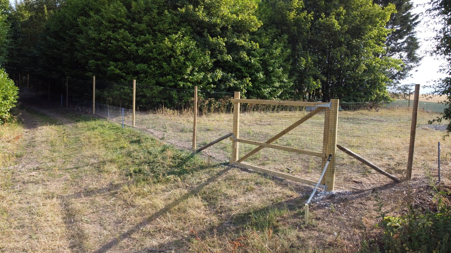 A wooden fence being constructed in a grassy field with trees in the background.