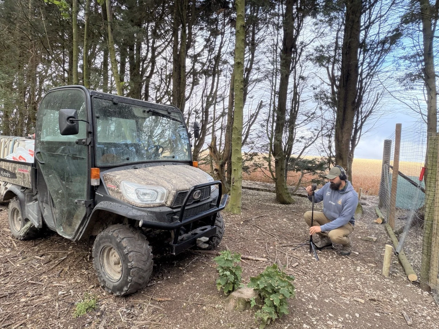 A person is kneeling on the ground in a wooded area with trees, using a tripod to set up equipment next to a small off-road utility vehicle. The person is wearing headphones and a cap, and there is a fence on the right side of the scene.