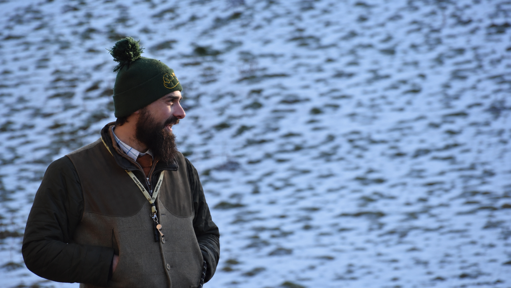 A man with a beard wearing a green beanie and a brown and gray jacket standing near a body of water, smiling.